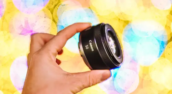 Dłoń trzyma obiektyw Canon 50mm, idealny do makro. Tło to kolorowe, rozmyte światła.